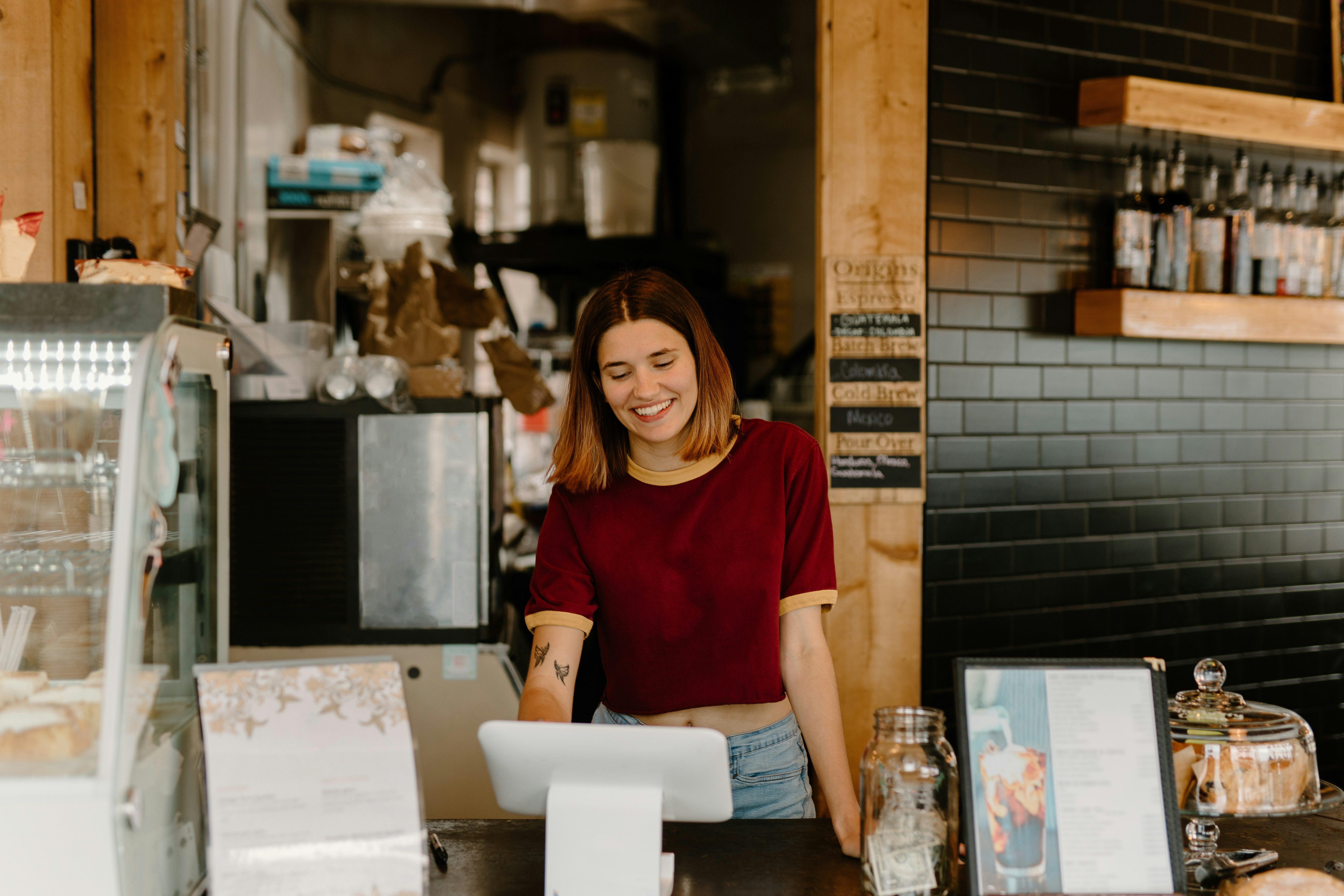 A smiling barista in a red shirt standing behind the counter of a cozy coffee shop, surrounded by baked goods, a menu, and a tip jar, creating a welcoming atmosphere.